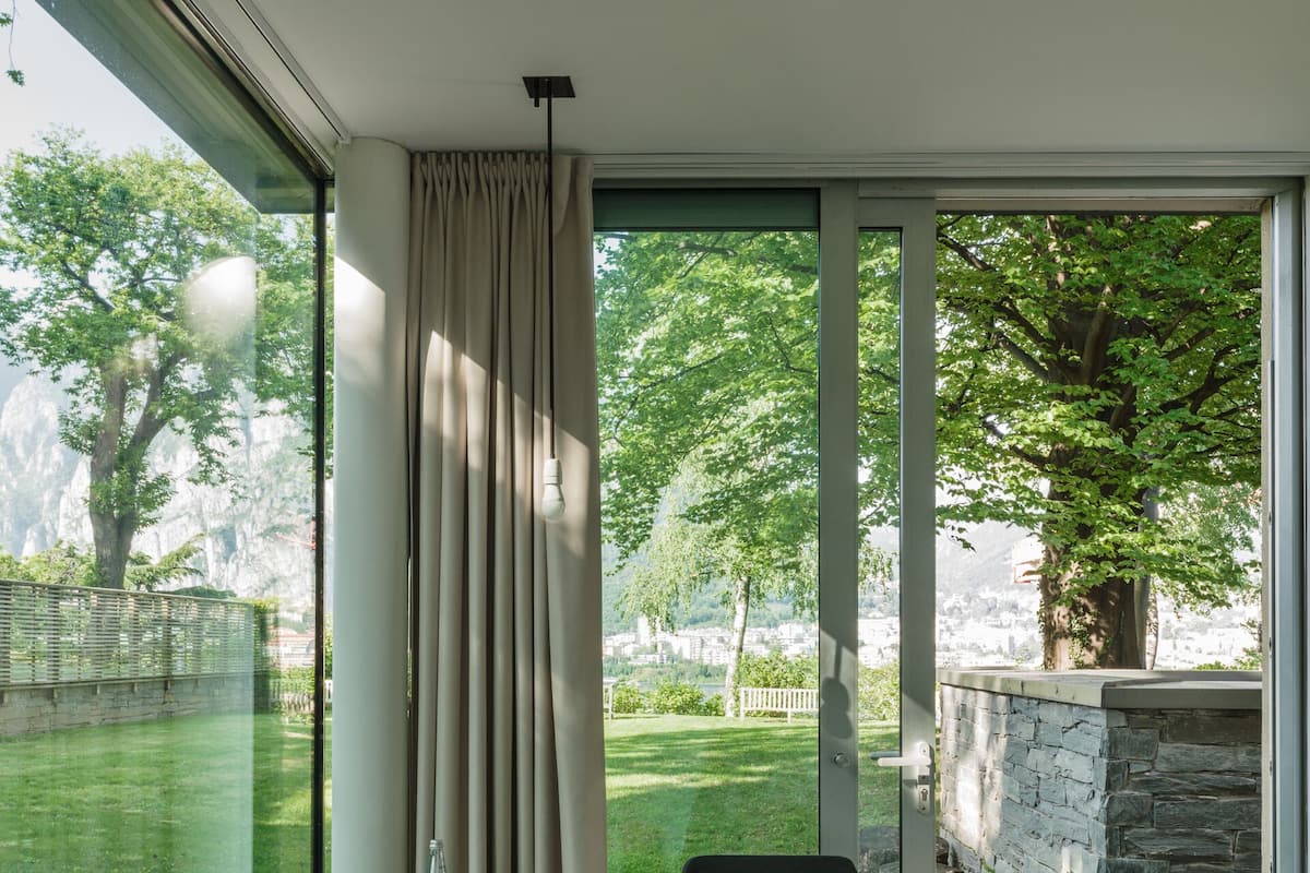 Une chambre moderne dotée d’une grande porte-fenêtre coulissante donnant sur un jardin. À l’intérieur, on trouve une table ronde en bois et deux chaises noires.