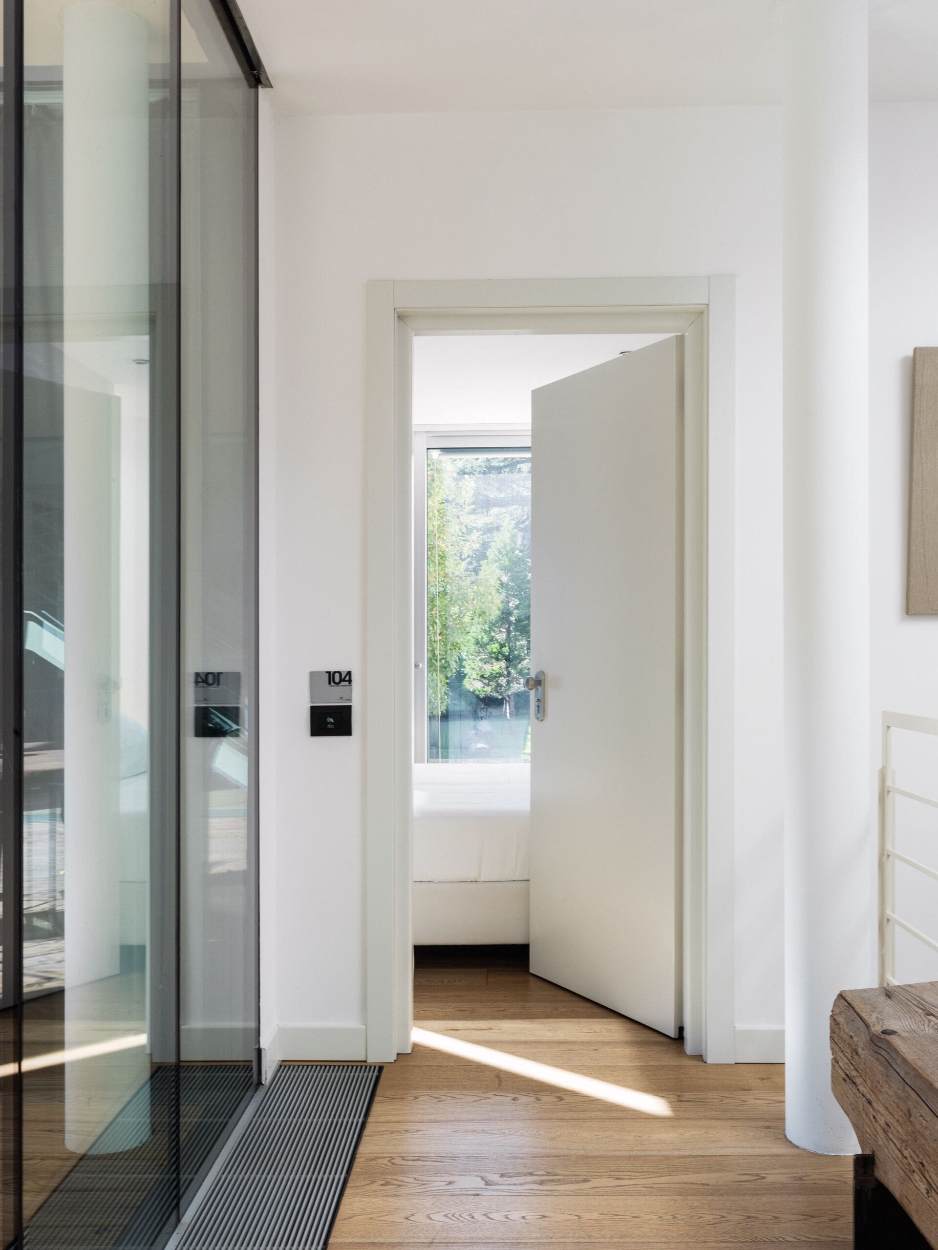 Un corridoio moderno con una porta in vetro, una panca in legno e vista su spazi verdi.
