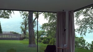 A bedroom with a large bed, a wooden nightstand, a chair, and a view of a lake and trees through floor-to-ceiling windows. - Casa sull'Albero (Malgrate)
