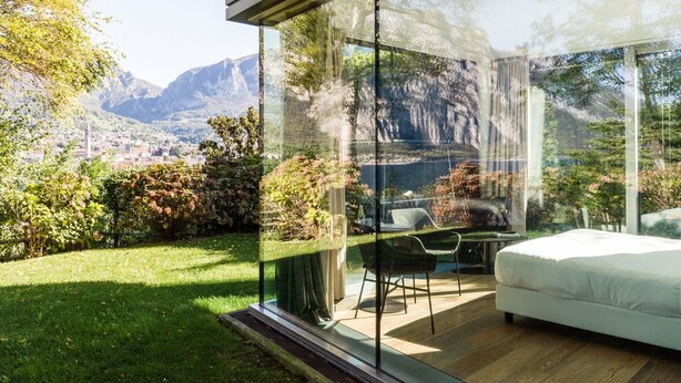 A modern bedroom with a glass wall, a bed, and a chair, overlooking a mountainous landscape. - Casa sull'Albero (Malgrate)