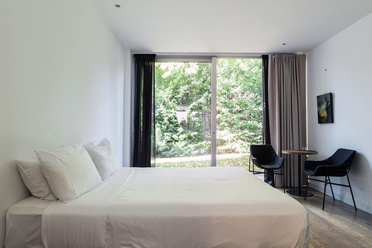 Une chambre d’hôtel moderne avec un grand lit, une petite table, deux chaises et une vue sur la verdure.
