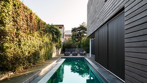 A modern outdoor pool area with a wooden deck, a stone wall, and a dark building facade. - Casa sull'Albero (Malgrate)