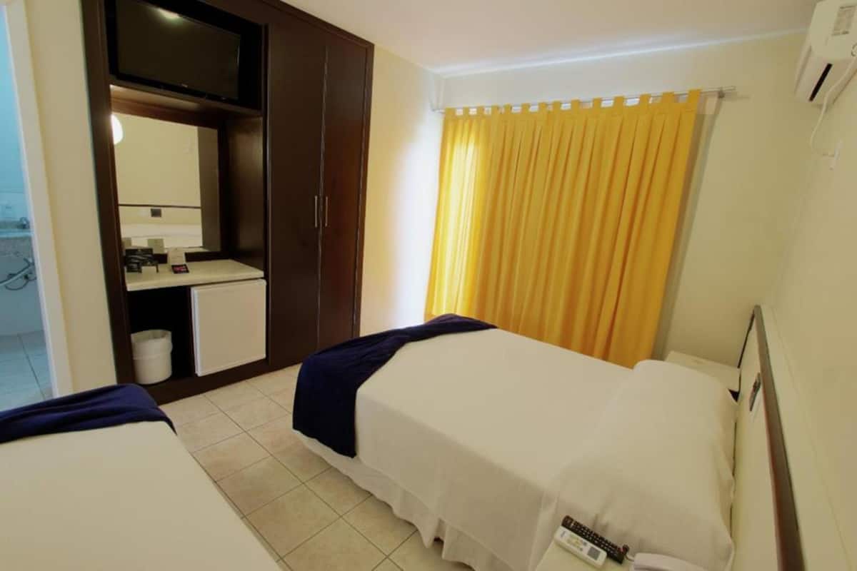 Habitación de hotel con dos camas, un escritorio pequeño con espejo, una cortina amarilla y una pared blanca.