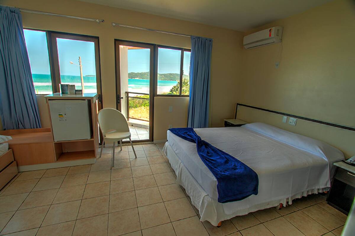 Habitación de hotel con cama, escritorio, silla y vista a la playa a través de amplias ventanas.