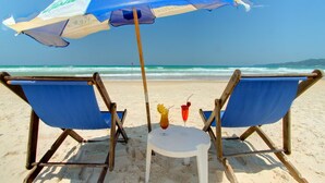 Two beach chairs with a blue umbrella and a small table with drinks. - Hotel Atlântico (Bombinhas)