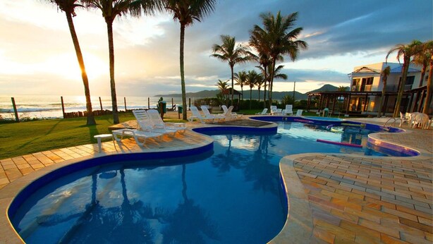 A swimming pool with lounge chairs, palm trees, and a view of the ocean at sunset. - Hotel Atlântico (Bombinhas)