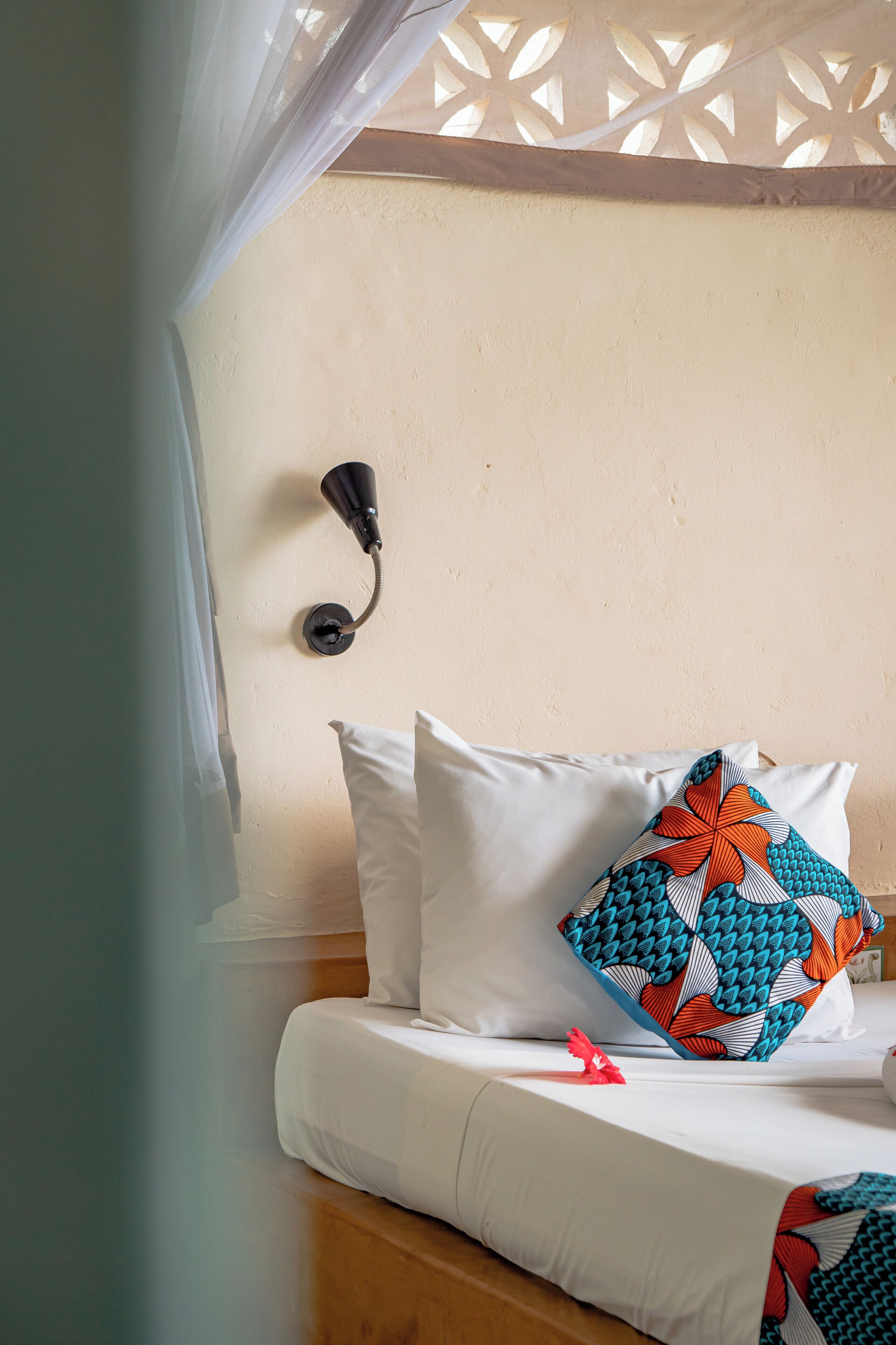 Un dormitorio con cama, ropa de cama blanca, un cojín decorativo, una lámpara de pared y una ventana con cortina translúcida.
