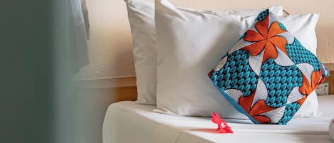 Un dormitorio con cama, ropa de cama blanca, un cojín decorativo, una lámpara de pared y una ventana con cortina translúcida.