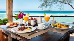 A breakfast table with plates of food, a pitcher, cups, and glasses of orange juice, overlooking a beach. - Sunshine Marine Lodge (Matemwe)