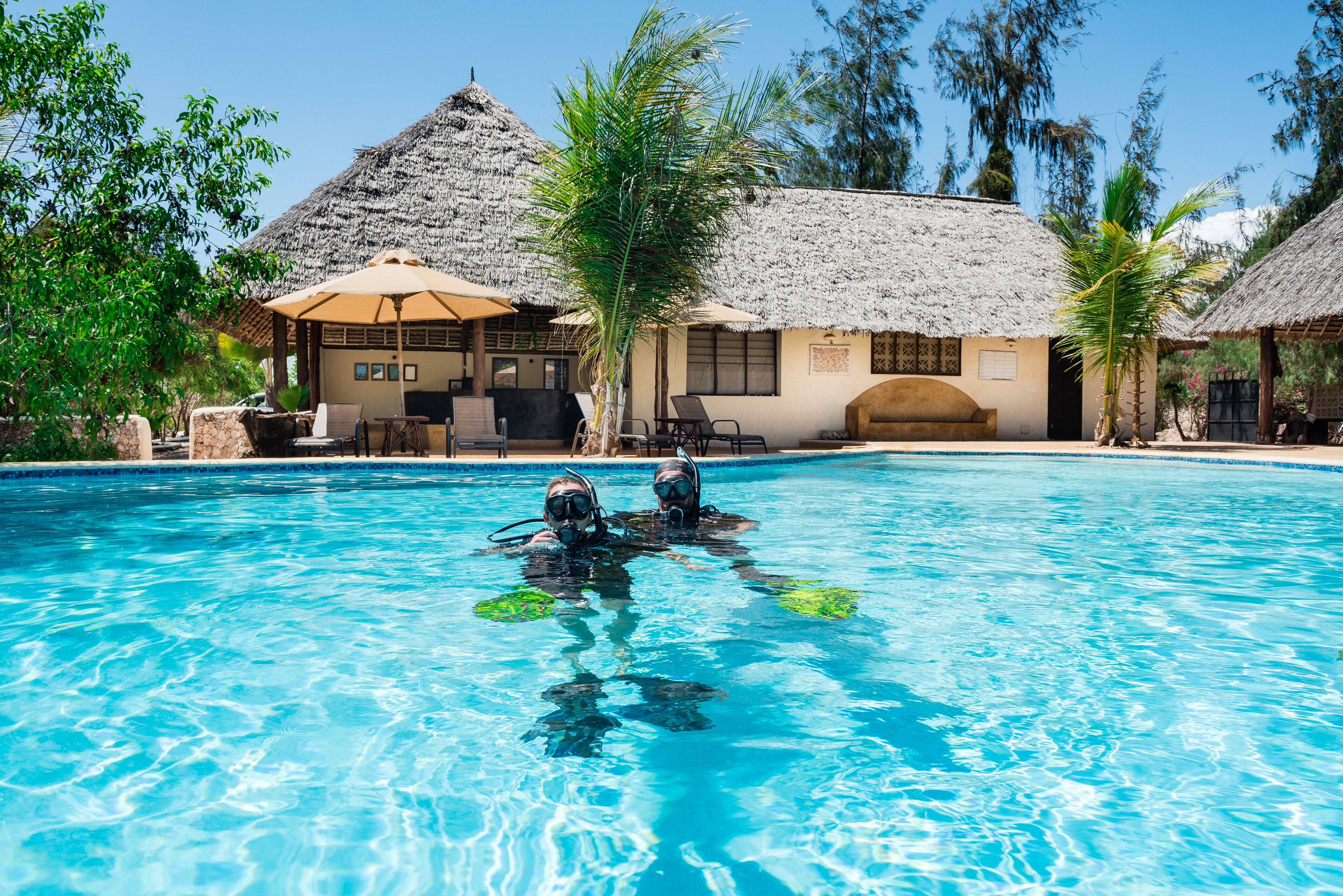 A clear blue pool with two people in scuba gear, surrounded by thatched-roof structures and palm trees.