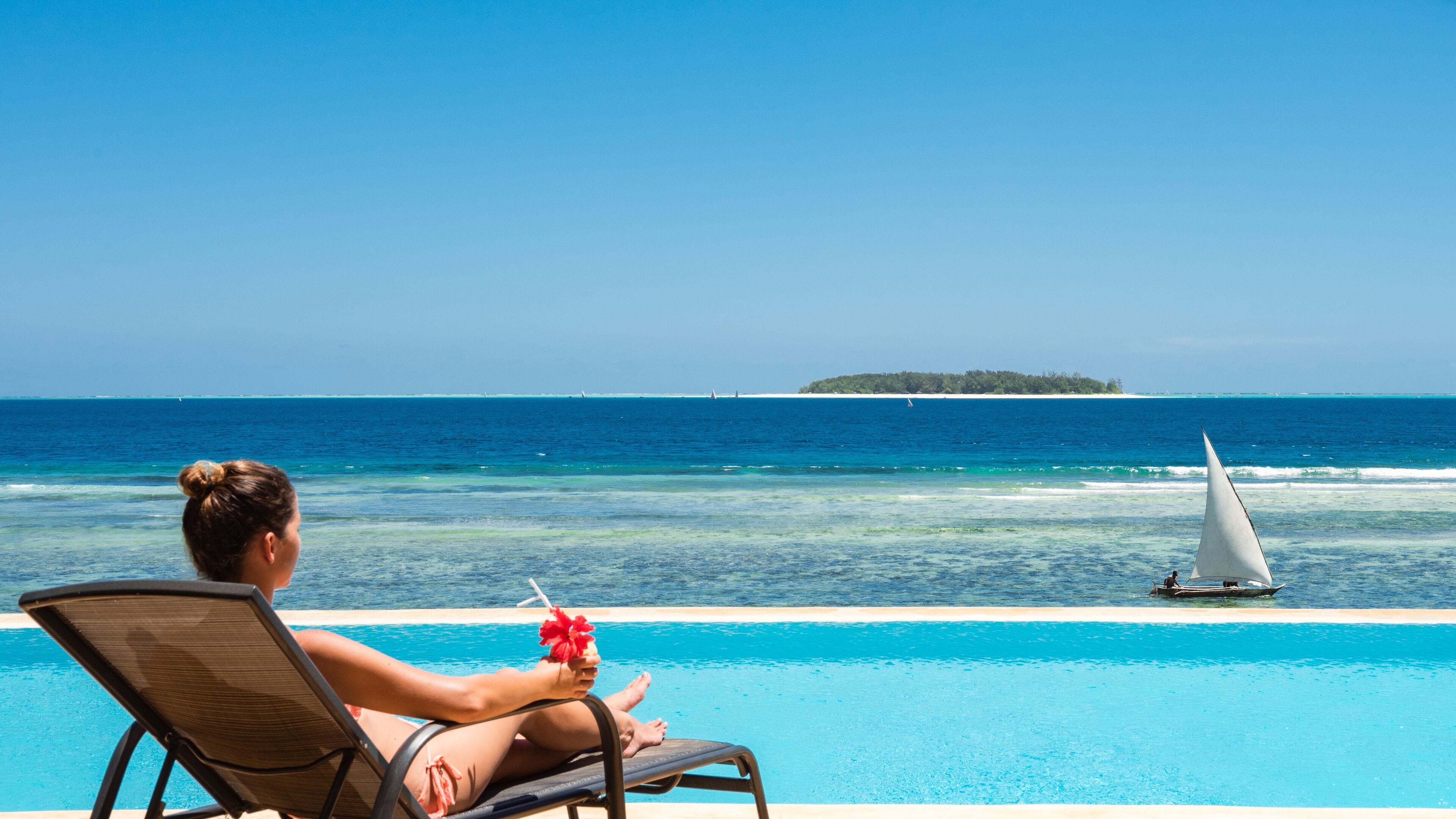 Une personne se détend au bord d’une piscine, avec vue sur l’océan et un voilier à l’horizon.