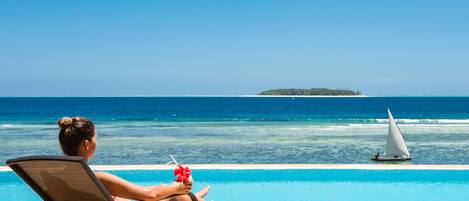 Une personne se détend au bord d’une piscine, avec vue sur l’océan et un voilier à l’horizon.