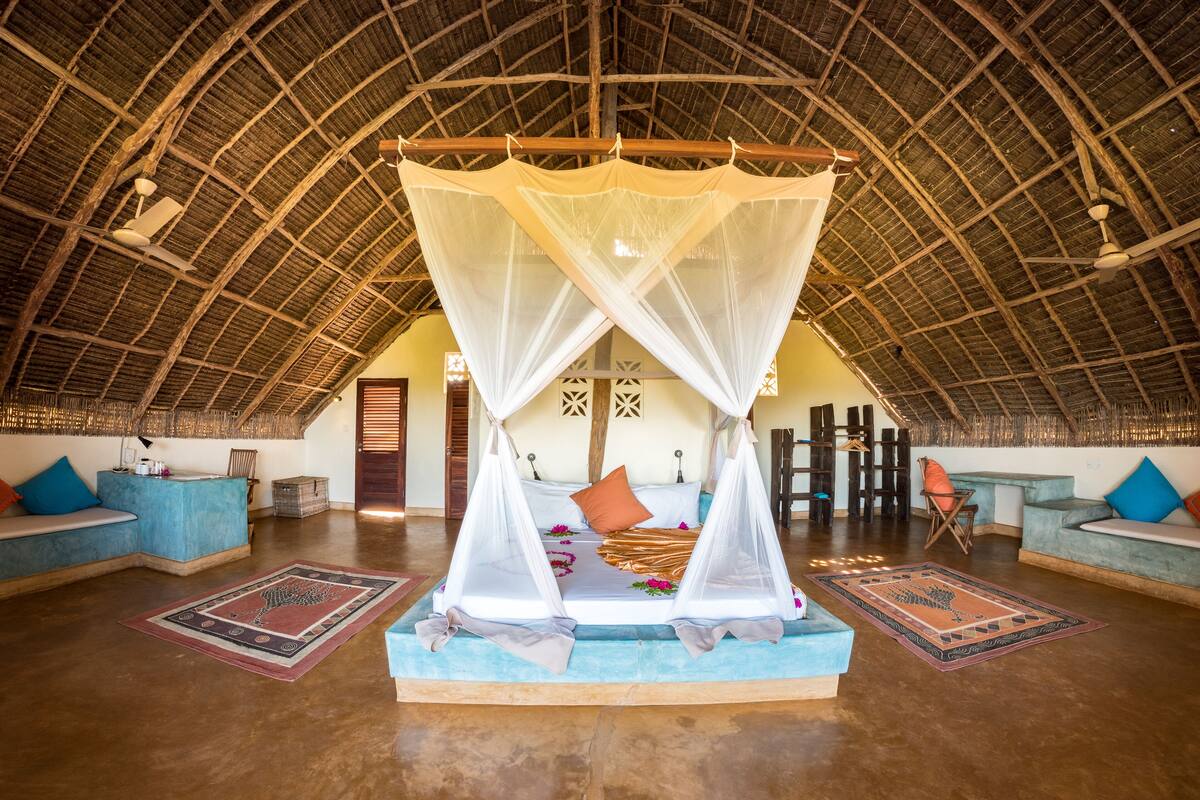 A spacious room with a high thatched ceiling, a central bed with a canopy, and two sofas with cushions.