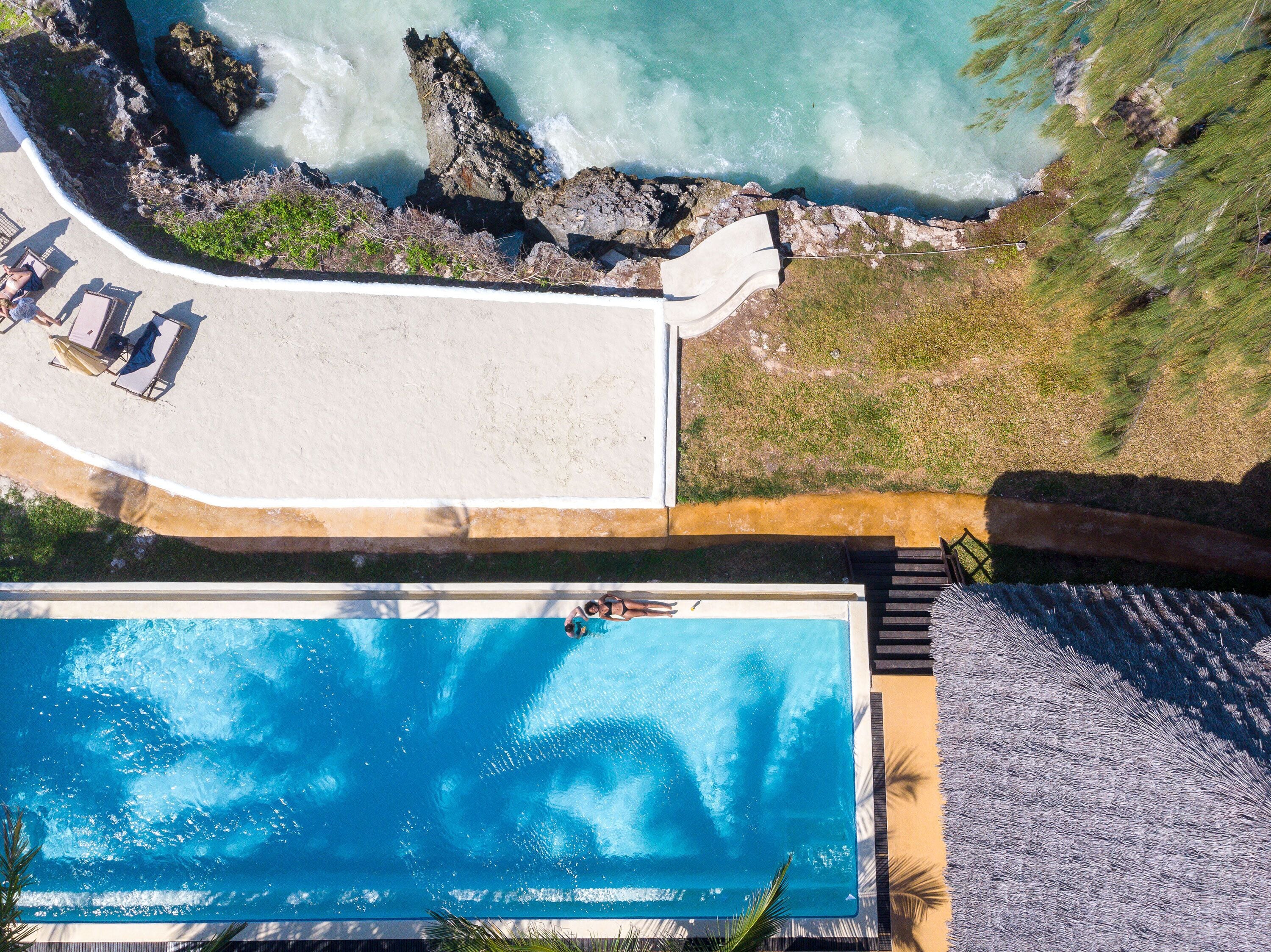 Vista aérea de un resort con piscina, zona de playa con sillones de descanso y costa rocosa.