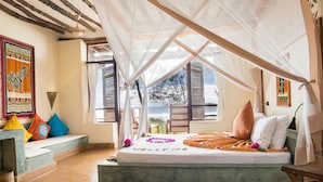 A bedroom with a bed under a canopy, a view of the outdoors, and a colorful wall decoration. - Sunshine Marine Lodge (Matemwe)