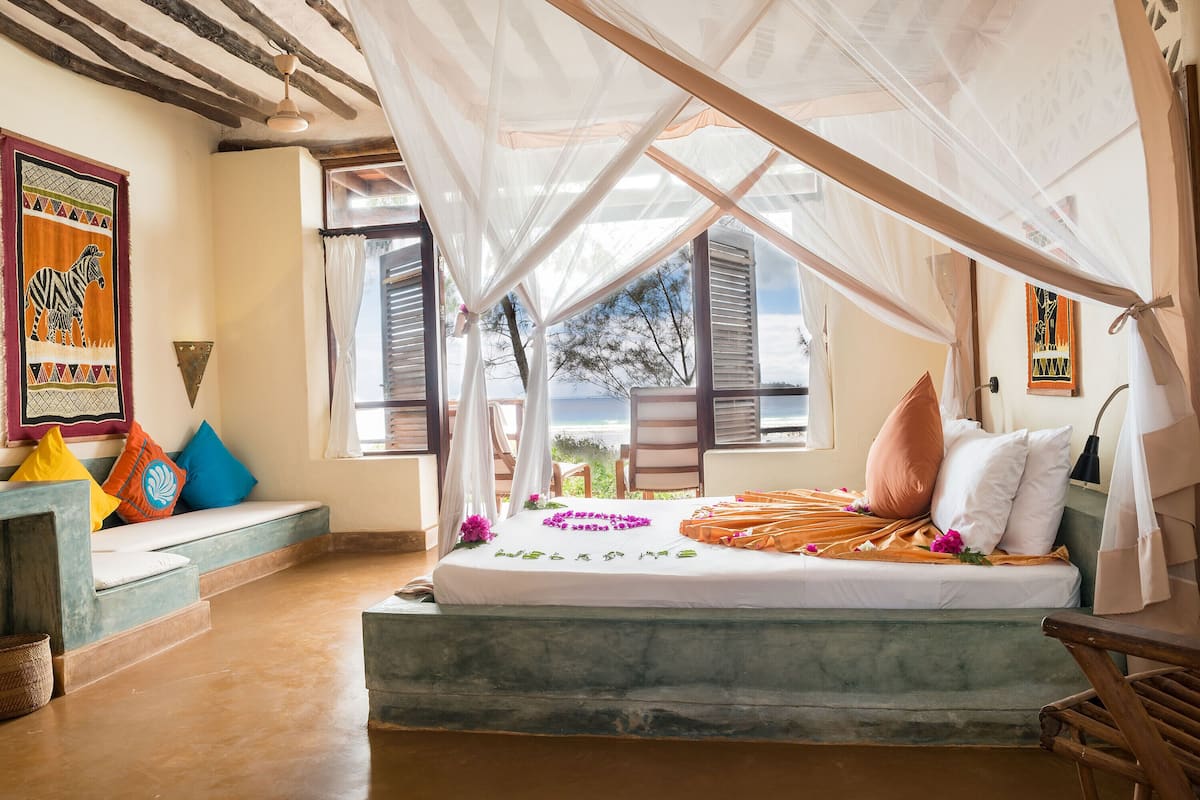 A bedroom with a bed under a canopy, a view of the outdoors, and a colorful wall decoration.