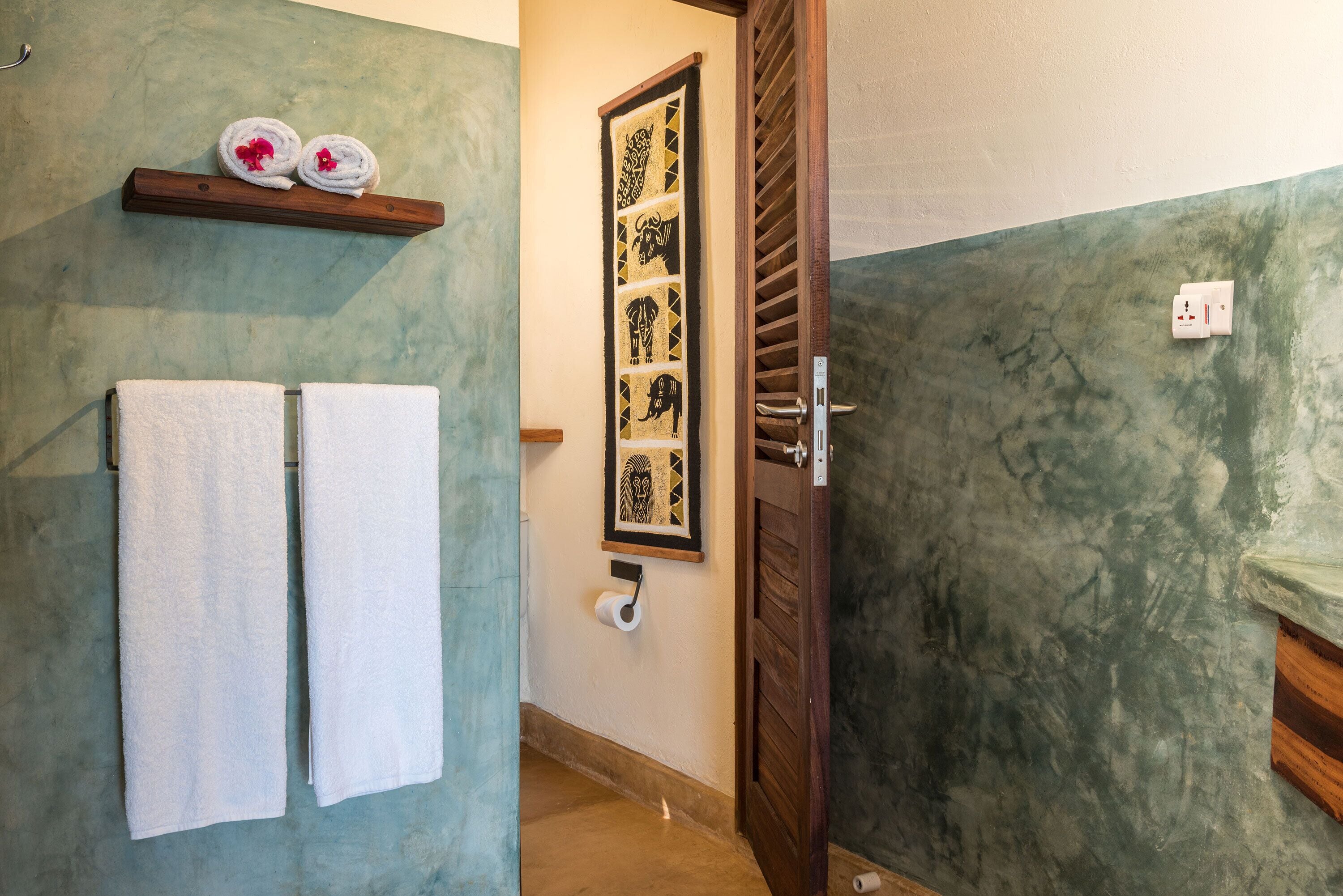 A bathroom with two white towels hanging on a rack, a wooden shelf with decorative towels, a framed artwork on the wall, and a door with a wooden frame.
