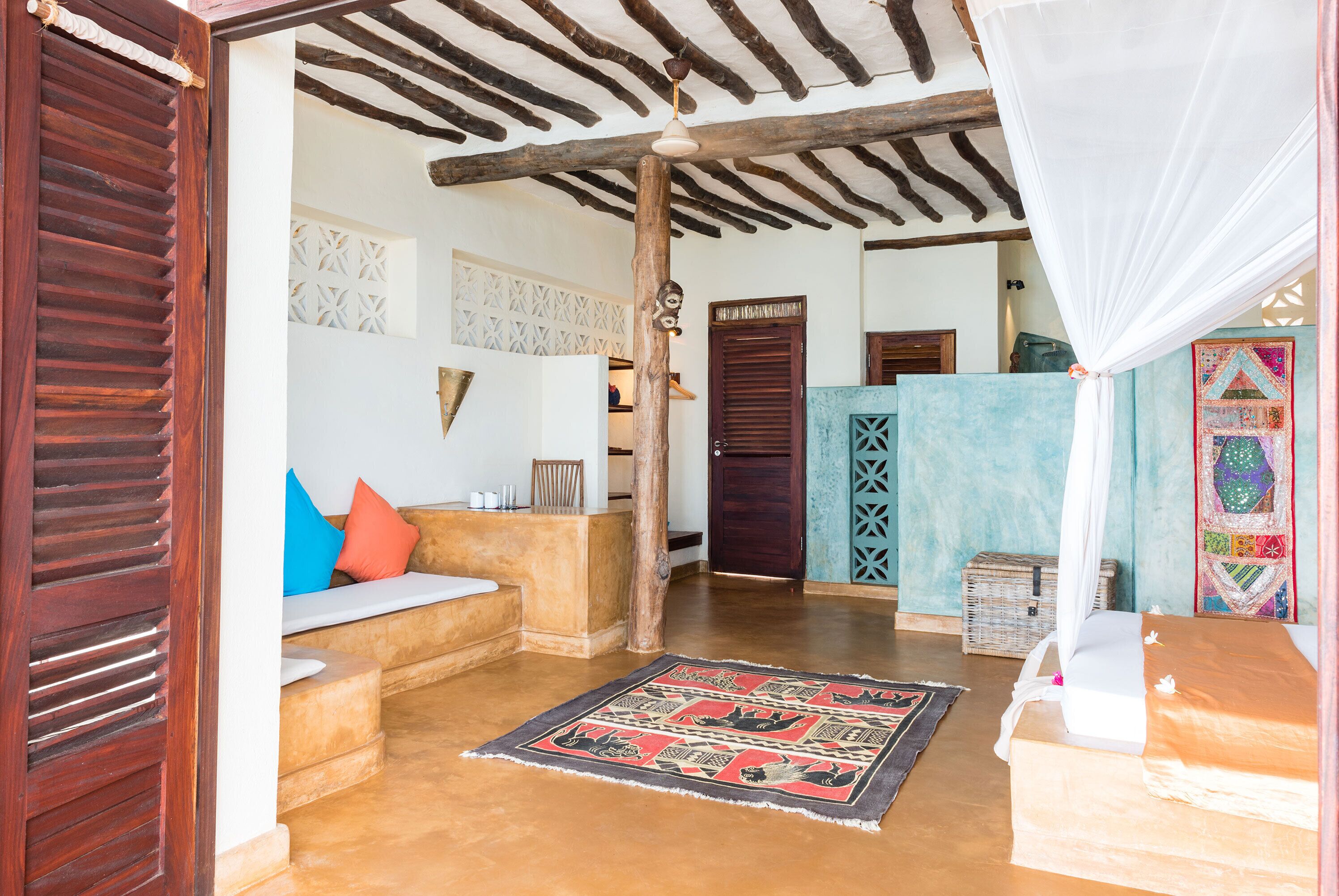 A room with a wooden ceiling, a bed with a white canopy, a colorful rug, and a wooden bench with cushions.