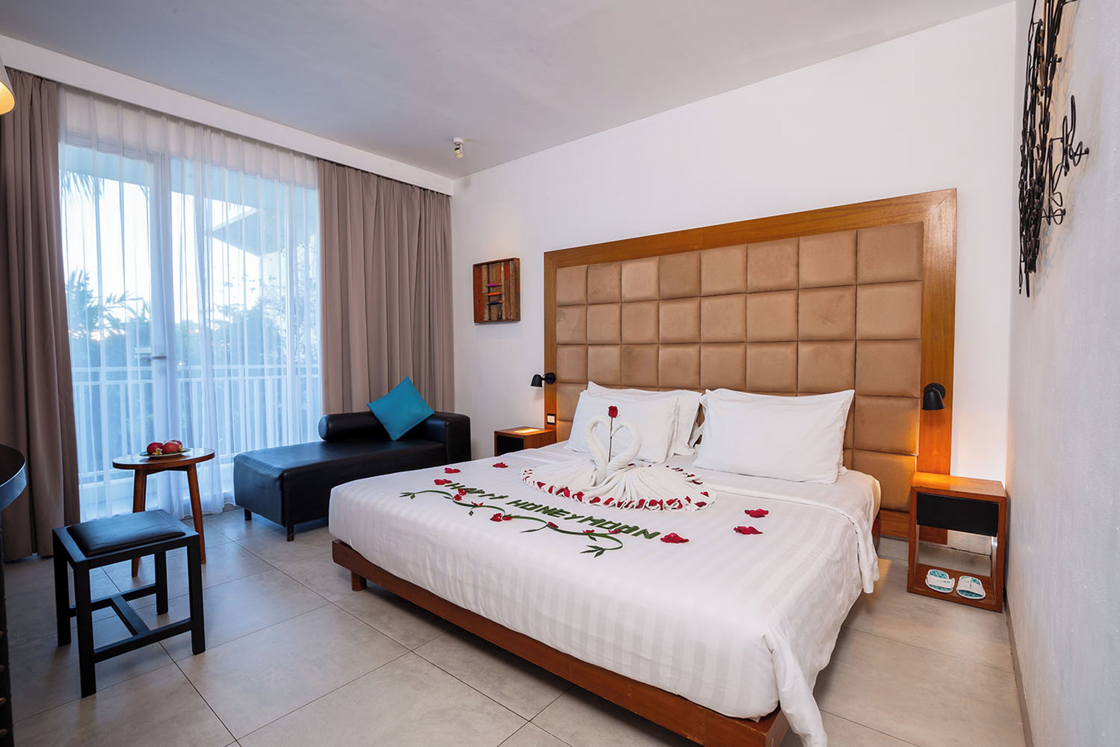 A hotel room with a large bed, a small table, a sofa, and a balcony with curtains.