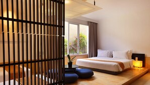 Una habitación de hotel moderna con una cama grande, una plataforma de madera, una mesita con una planta y vistas al verdor exterior.