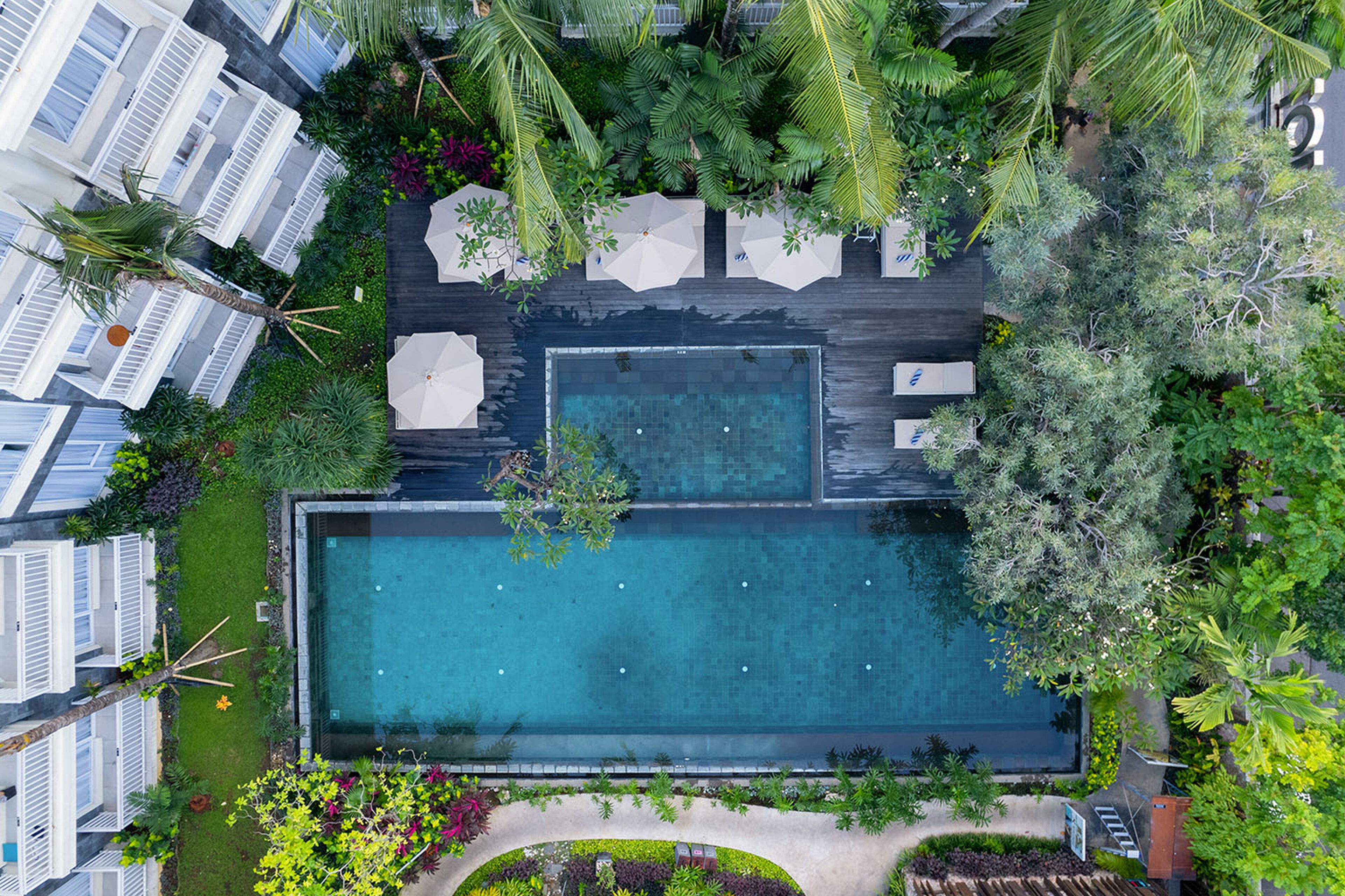 Vista aérea de un hotel con una piscina rectangular rodeada de exuberante vegetación, muebles de exterior blancos y un jardín bien cuidado.
