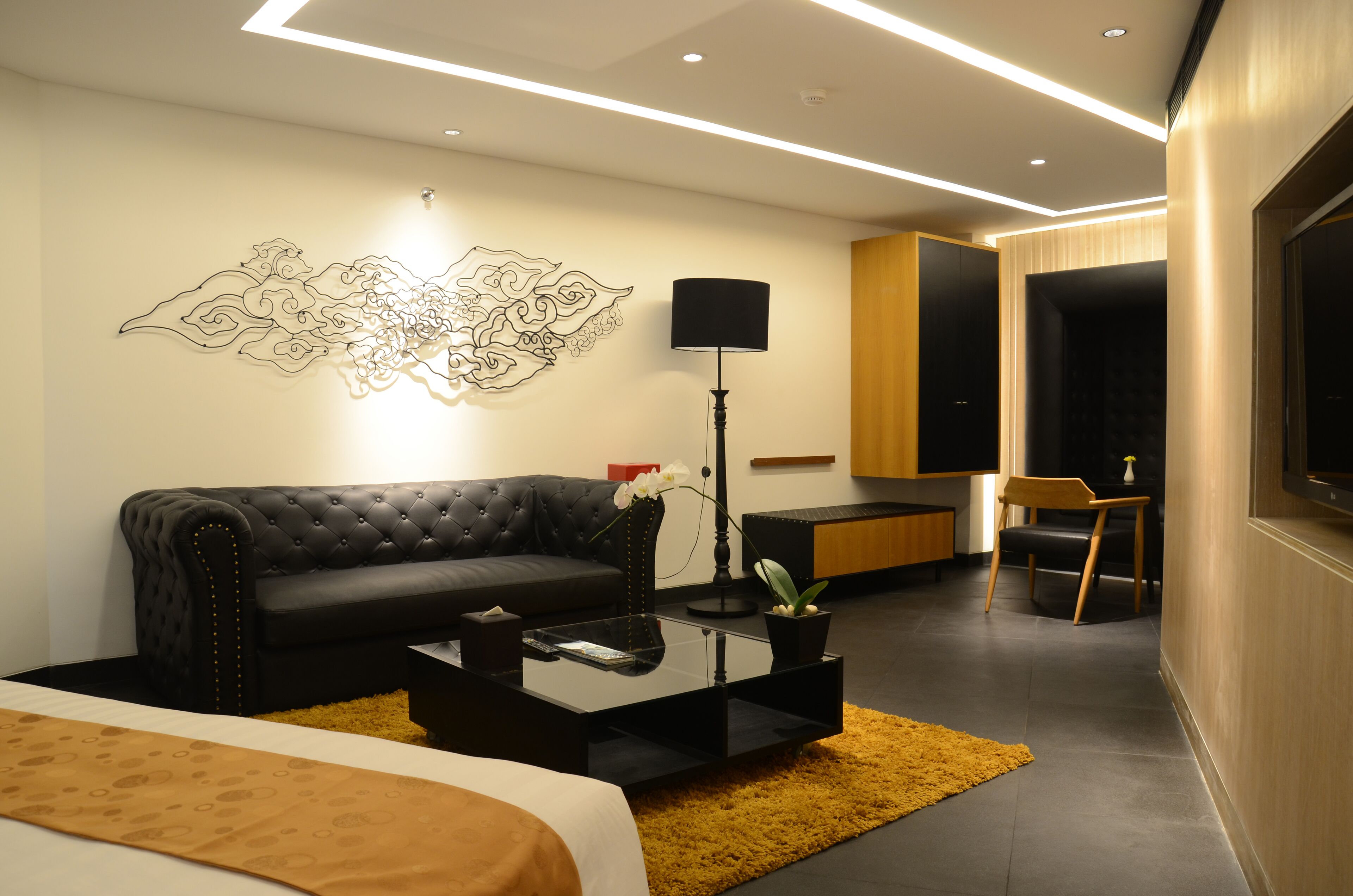 A modern hotel room with a black leather sofa, a glass coffee table, a black floor lamp, and a decorative wall piece.