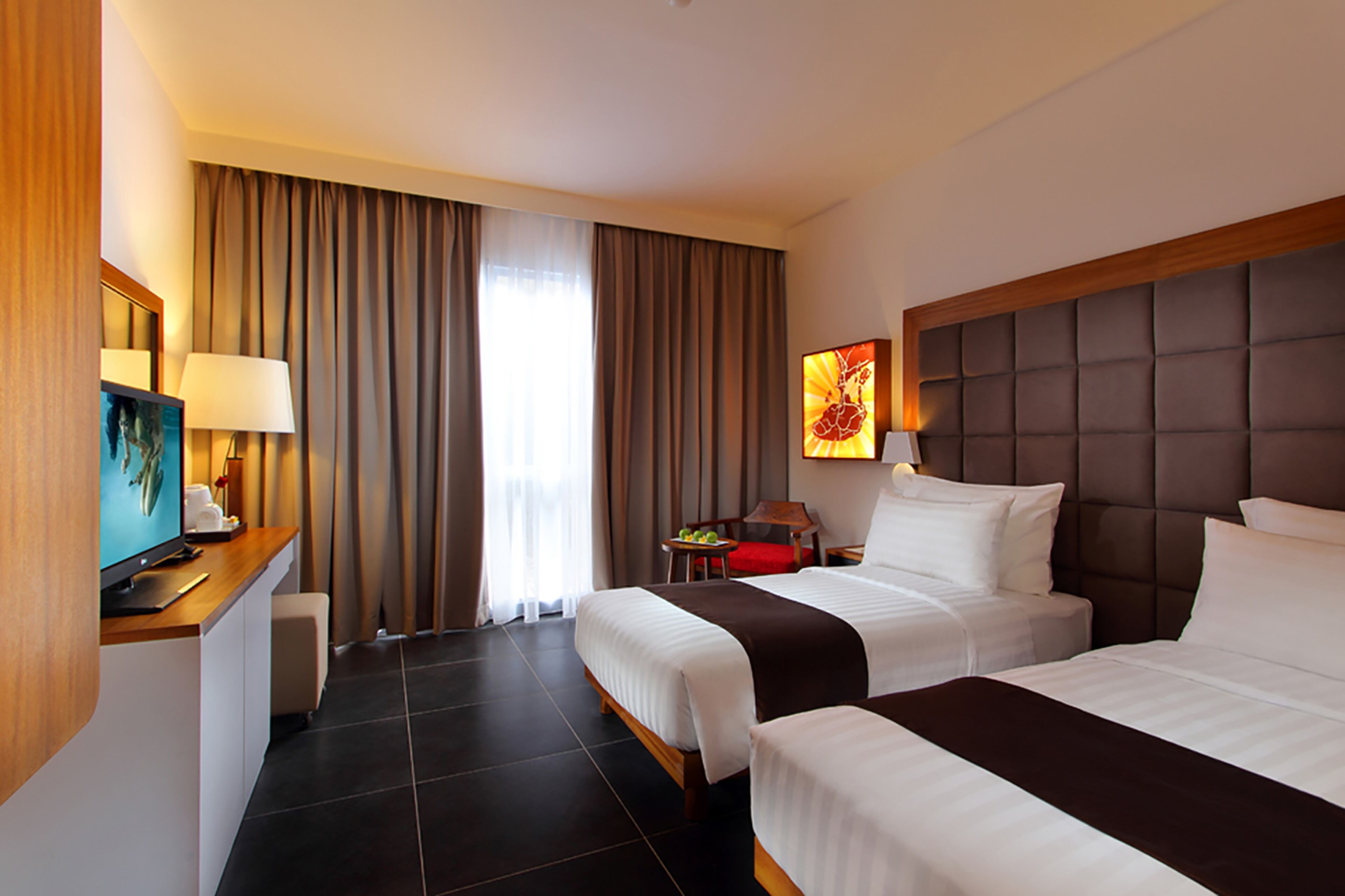 A hotel room with two beds, a desk with a computer, a chair, a small table with fruit, and a large window with curtains.
