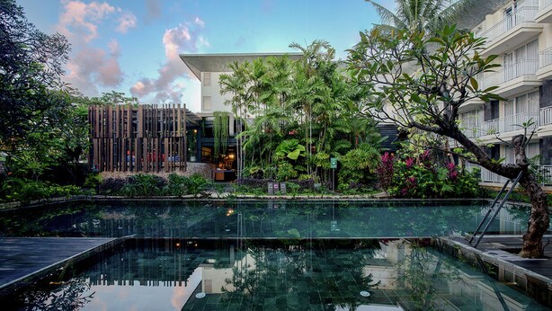 Un hotel moderno con piscina de fondo de cristal rodeado de exuberante vegetación y un edificio de varios pisos con balcones.