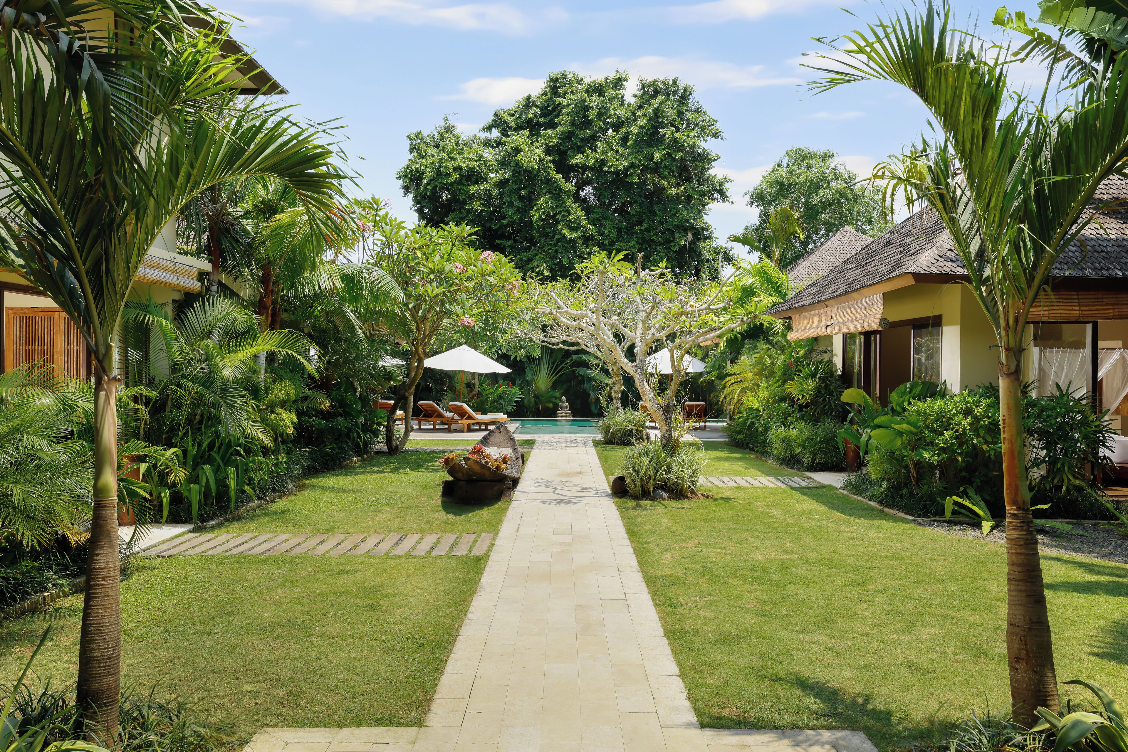 A well-maintained garden with a central pathway leading to a pool, surrounded by lush greenery and palm trees.