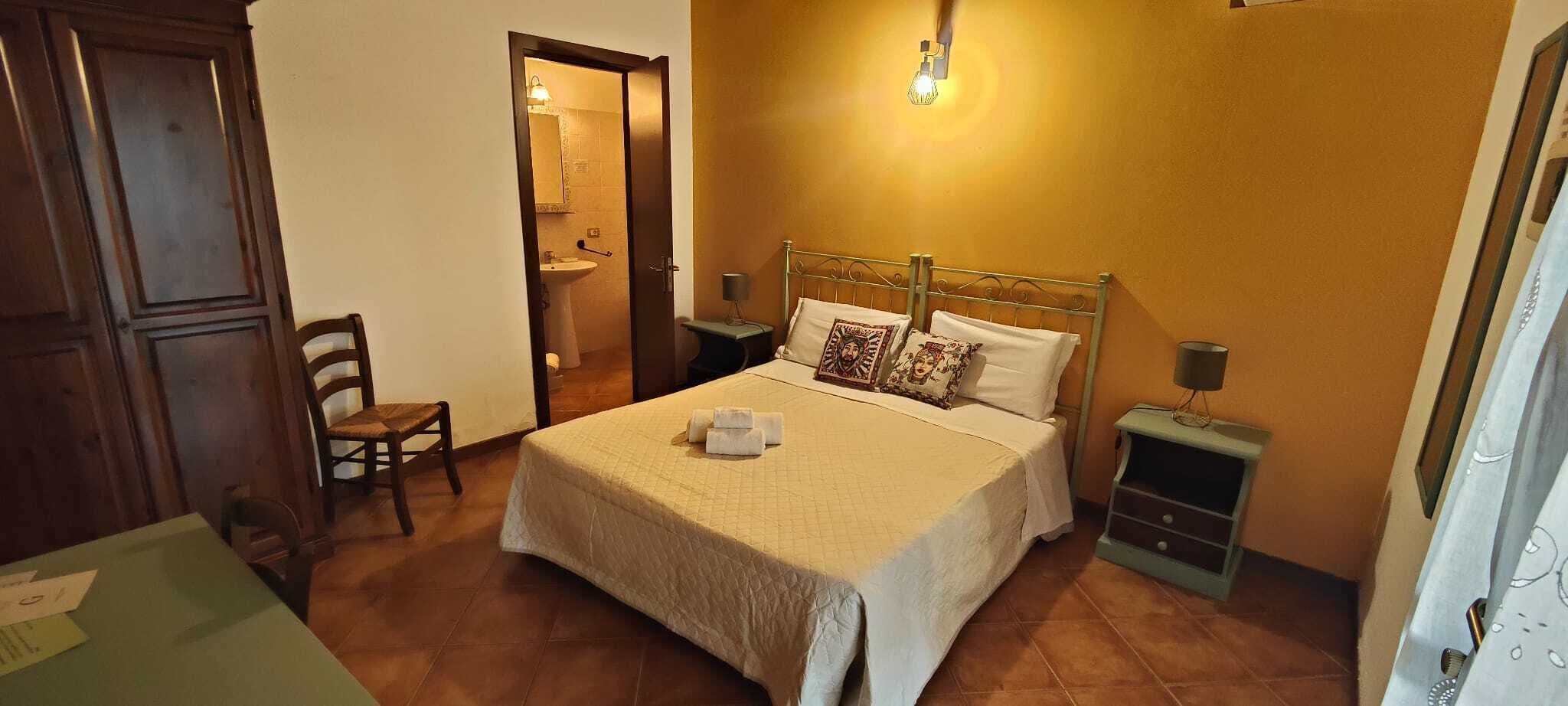 Een slaapkamer met een bed, een stoel, een nachtkastje en een badkamer zichtbaar door een geopende deur.