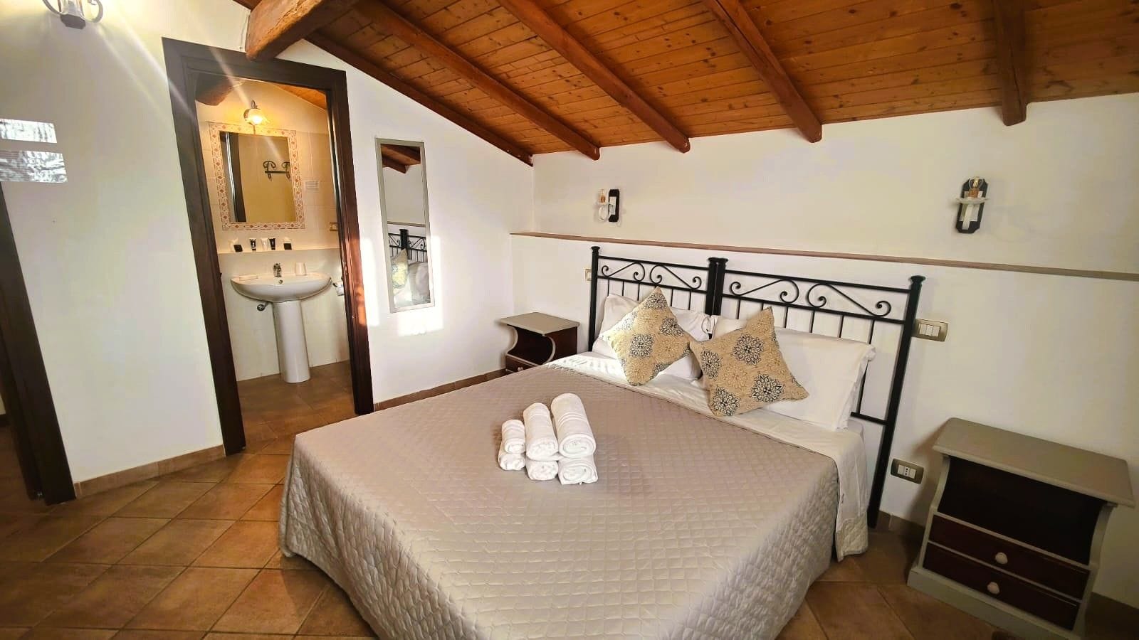 A bedroom with a bed, a bedside table, a nightstand, a mirror, and a wooden ceiling.