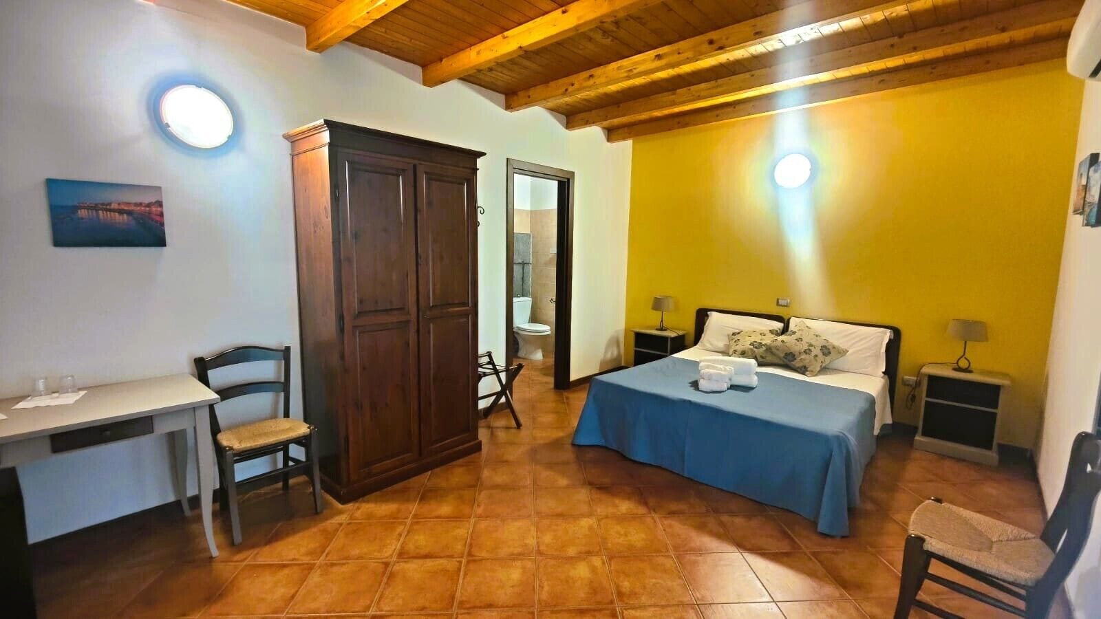 A bedroom with a bed, a desk, a chair, a wardrobe, and a bathroom visible through an open door.