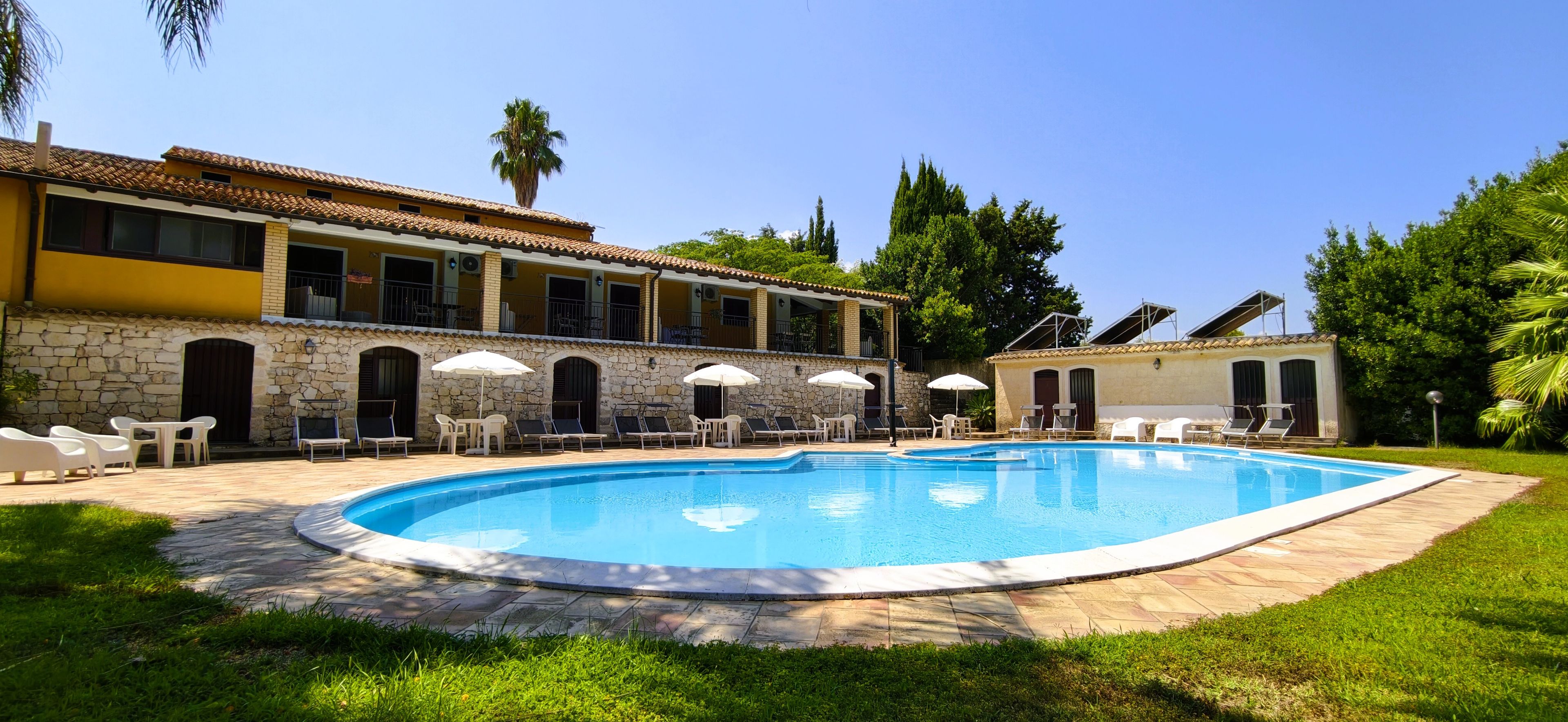A swimming pool with lounge chairs and umbrellas, surrounded by a stone wall and a building with multiple windows and balconies.