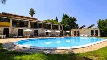 A swimming pool with lounge chairs and umbrellas, surrounded by a stone wall and a building with multiple windows and balconies.