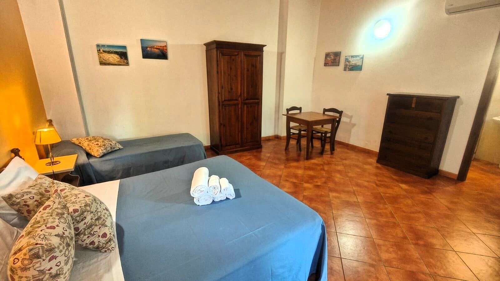 A room with two beds, a wooden wardrobe, a small table with chairs, and a lamp.