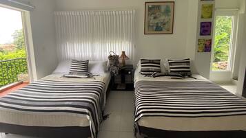 A room with two beds, each with striped bedding, a small table with a lamp, and a window with a view of greenery.