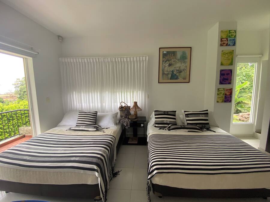 A room with two beds, each with striped bedding, a small table with a lamp, and a window with a view of greenery.