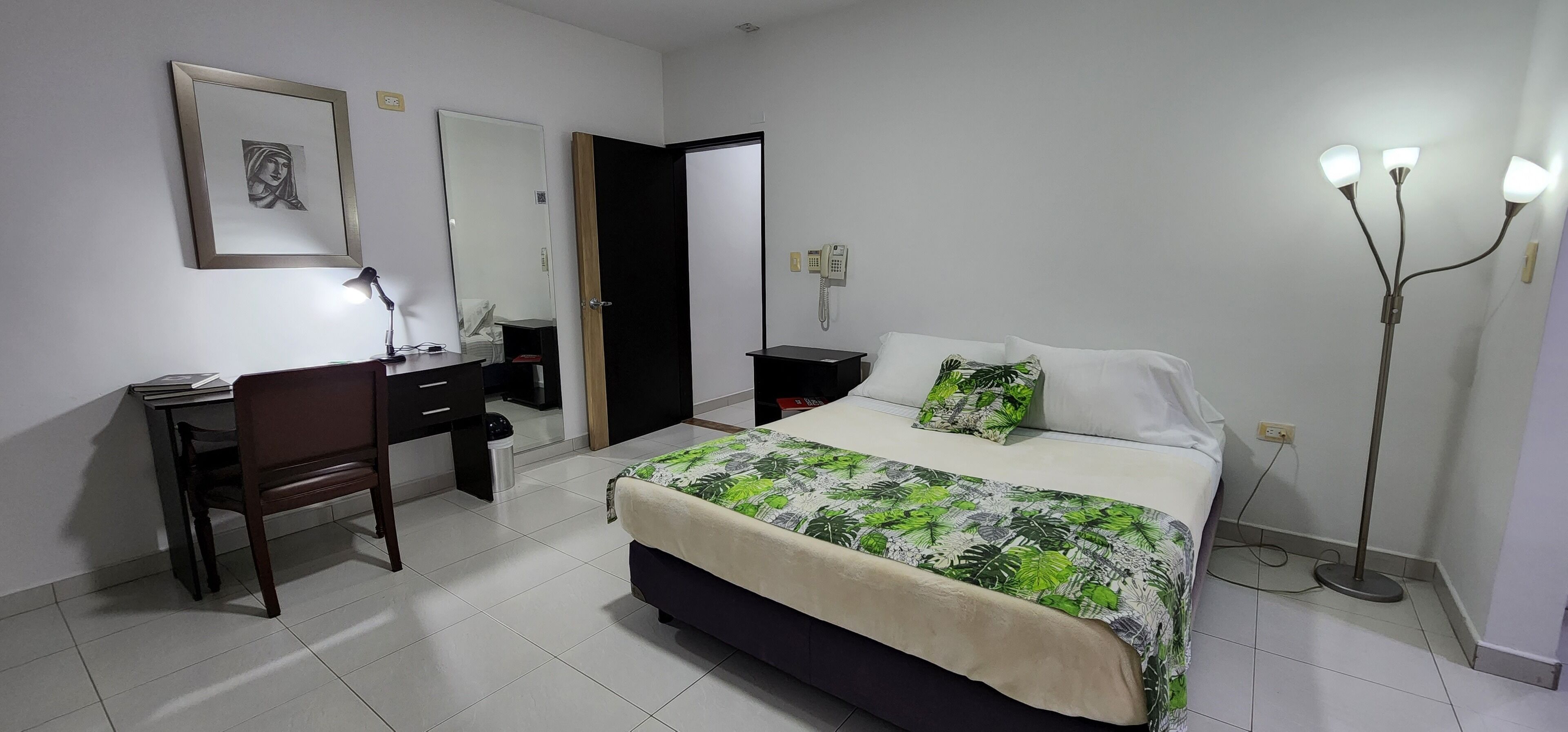 Habitació d'hotel amb llit, escriptori, cadira, taulell de nit i làmpada de peu.