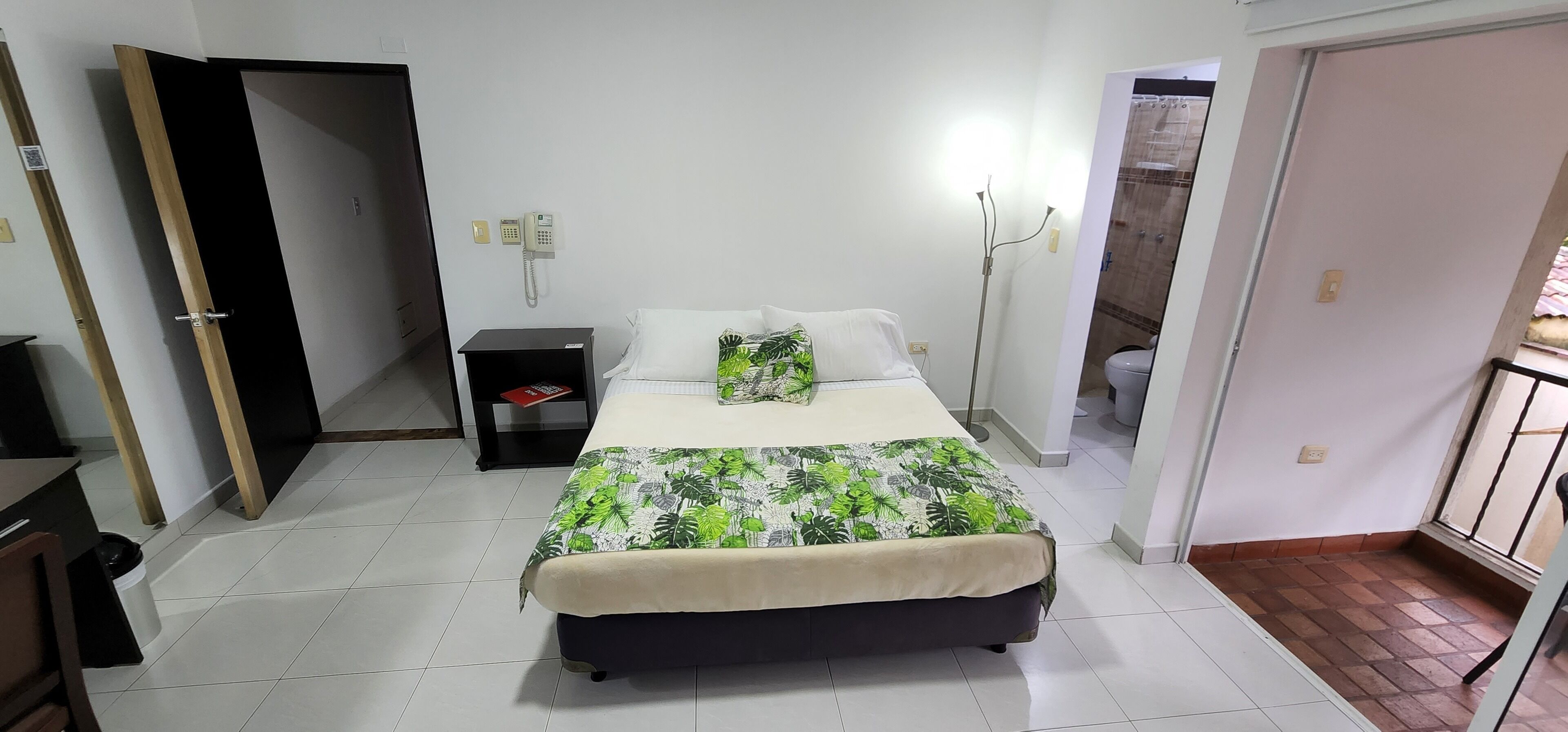 Un dormitorio con cama, mesita de noche y tocador. Hay una puerta que da a otra habitación y una ventana con vistas.