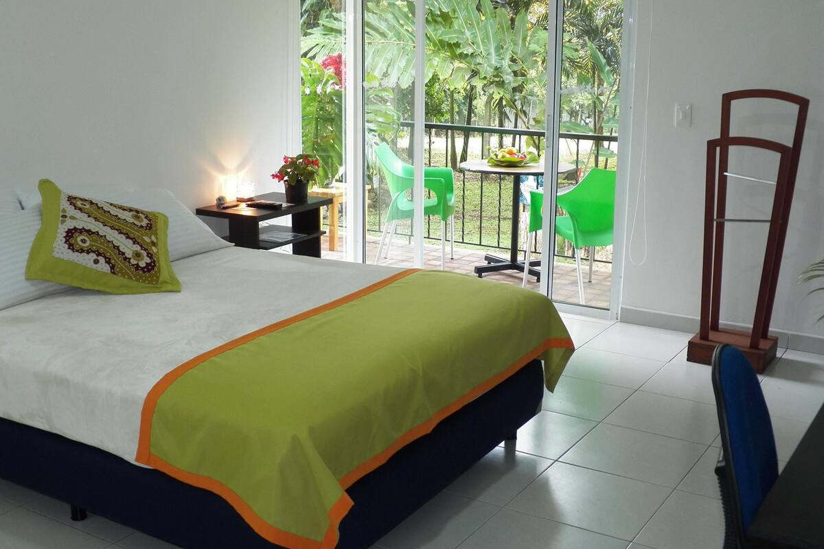 Une chambre avec un lit, une table de chevet, une table sur laquelle se trouve un panier de fruits, des chaises vertes et une vue sur la verdure à travers une porte vitrée.