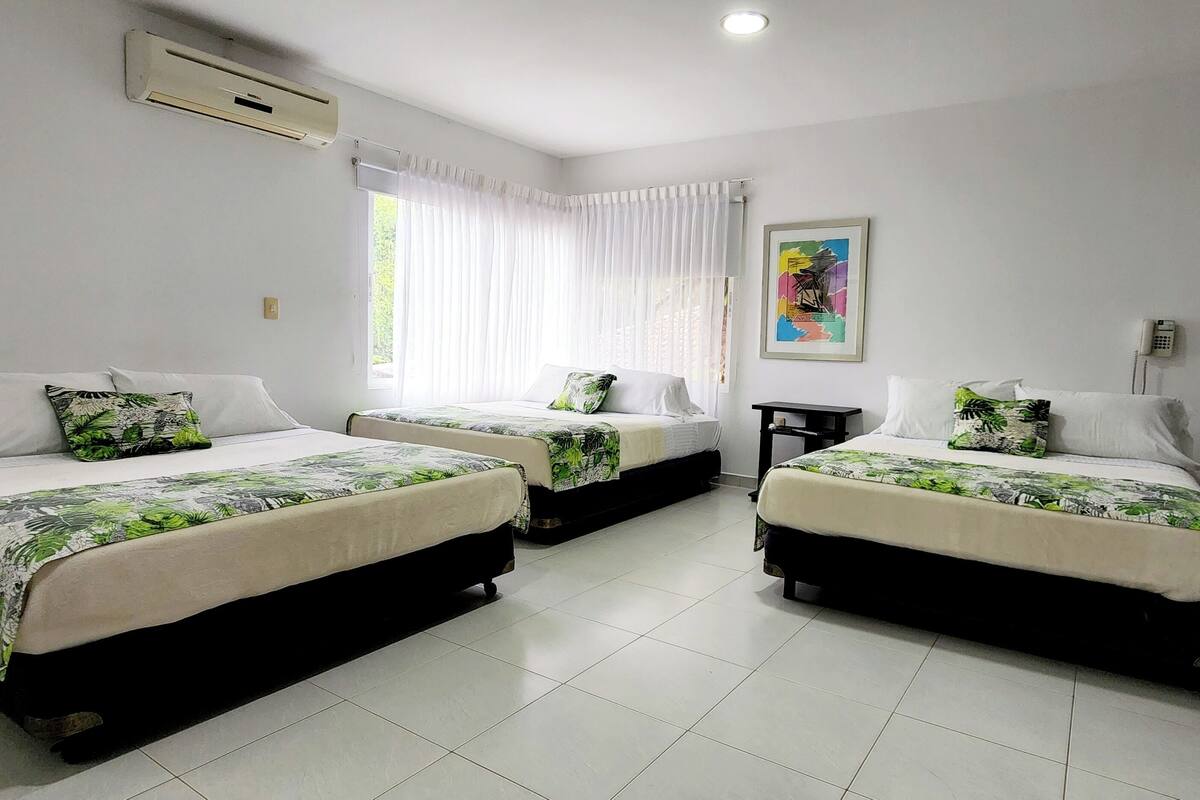 Une chambre d’hôtel avec trois lits simples, chacun agrémenté d’une couvre-lit aux motifs tropicaux, d’oreillers blancs et d’une petite table de chevet. La chambre dispose d’une fenêtre avec des rideaux légers, d’un climatiseur fixé au mur et d’un tableau accroché au mur.