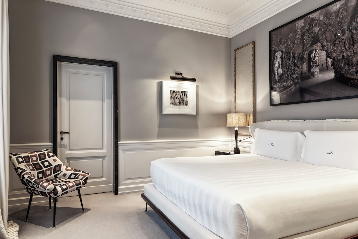 A bedroom with a large bed, a chair, a door, a framed picture, and a lamp.