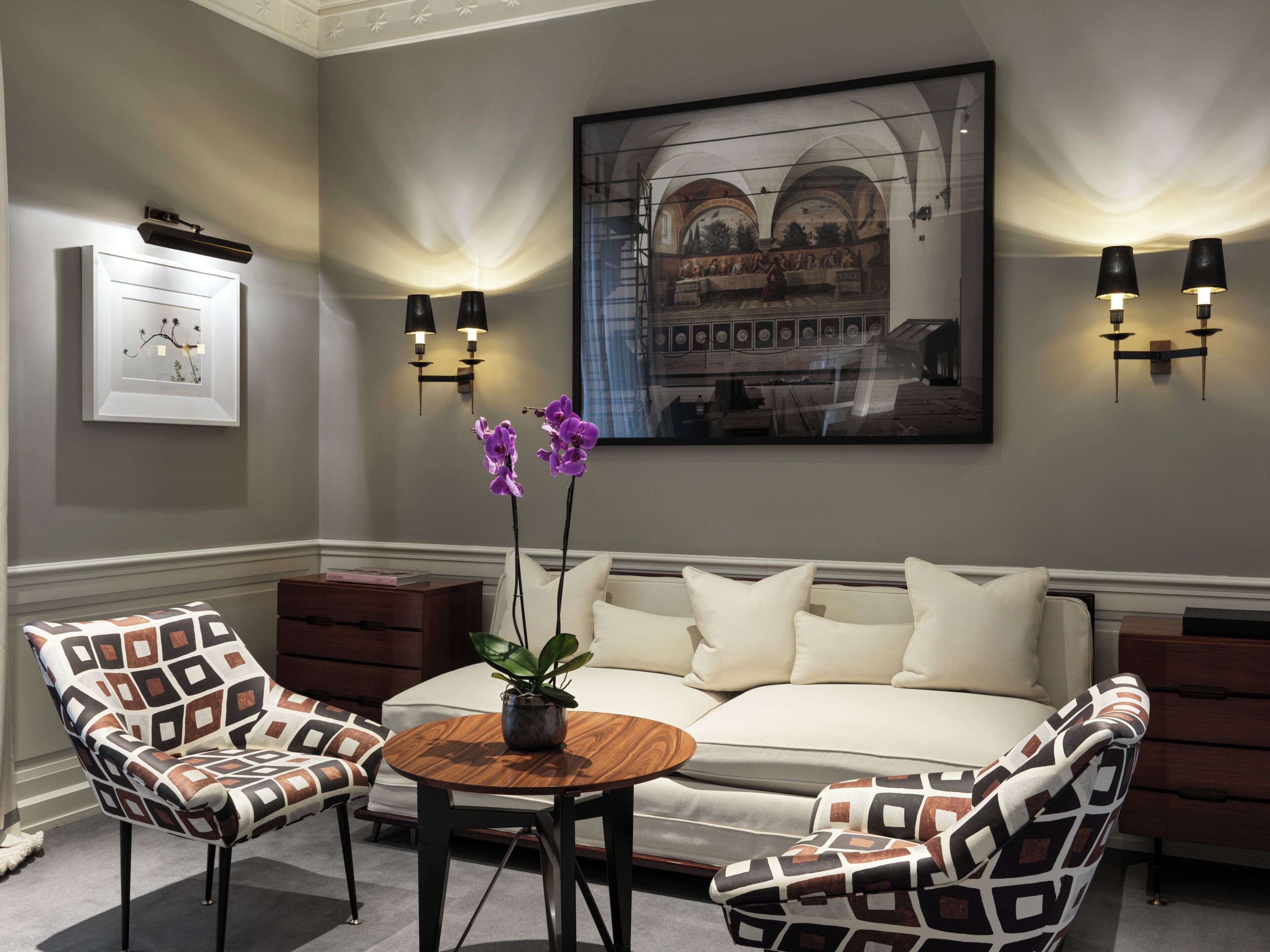 A room with a sofa, two patterned armchairs, a small wooden table with a plant, and wall-mounted lighting.