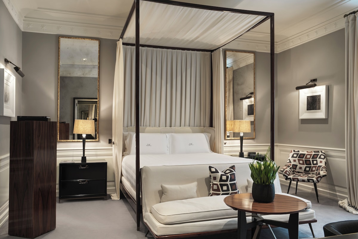 A four-poster canopy bed with a bedside table, a chair with a patterned cushion, and a small table with a plant.