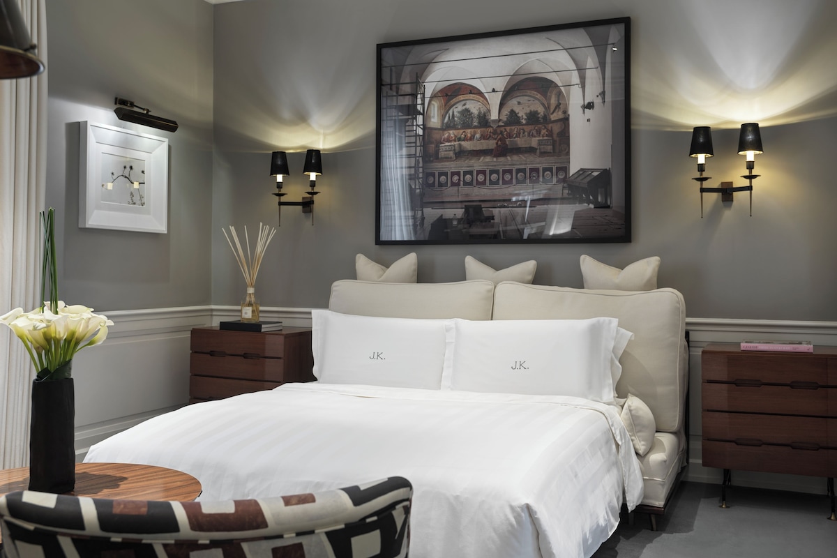 A bedroom with a large bed, two bedside tables, a chair, a vase with flowers, and wall-mounted lighting.