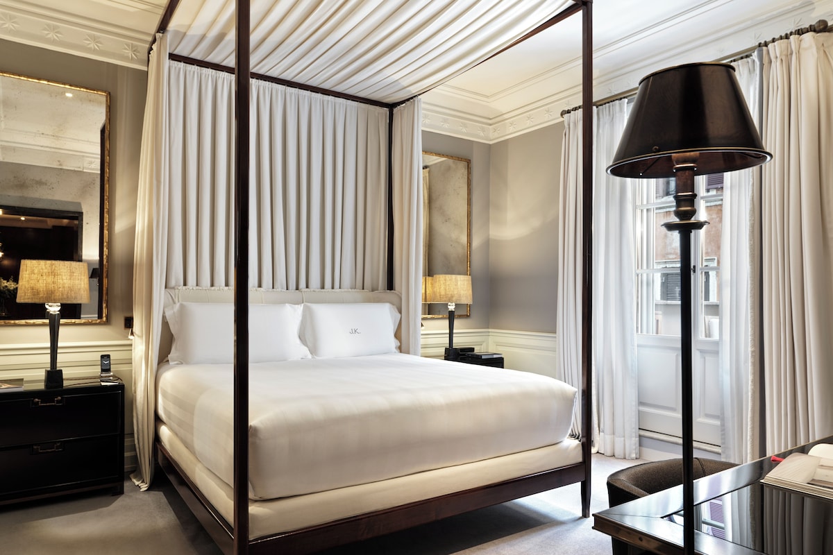 A four-poster bed with white linens, bedside lamps, a nightstand, and a desk with a chair.