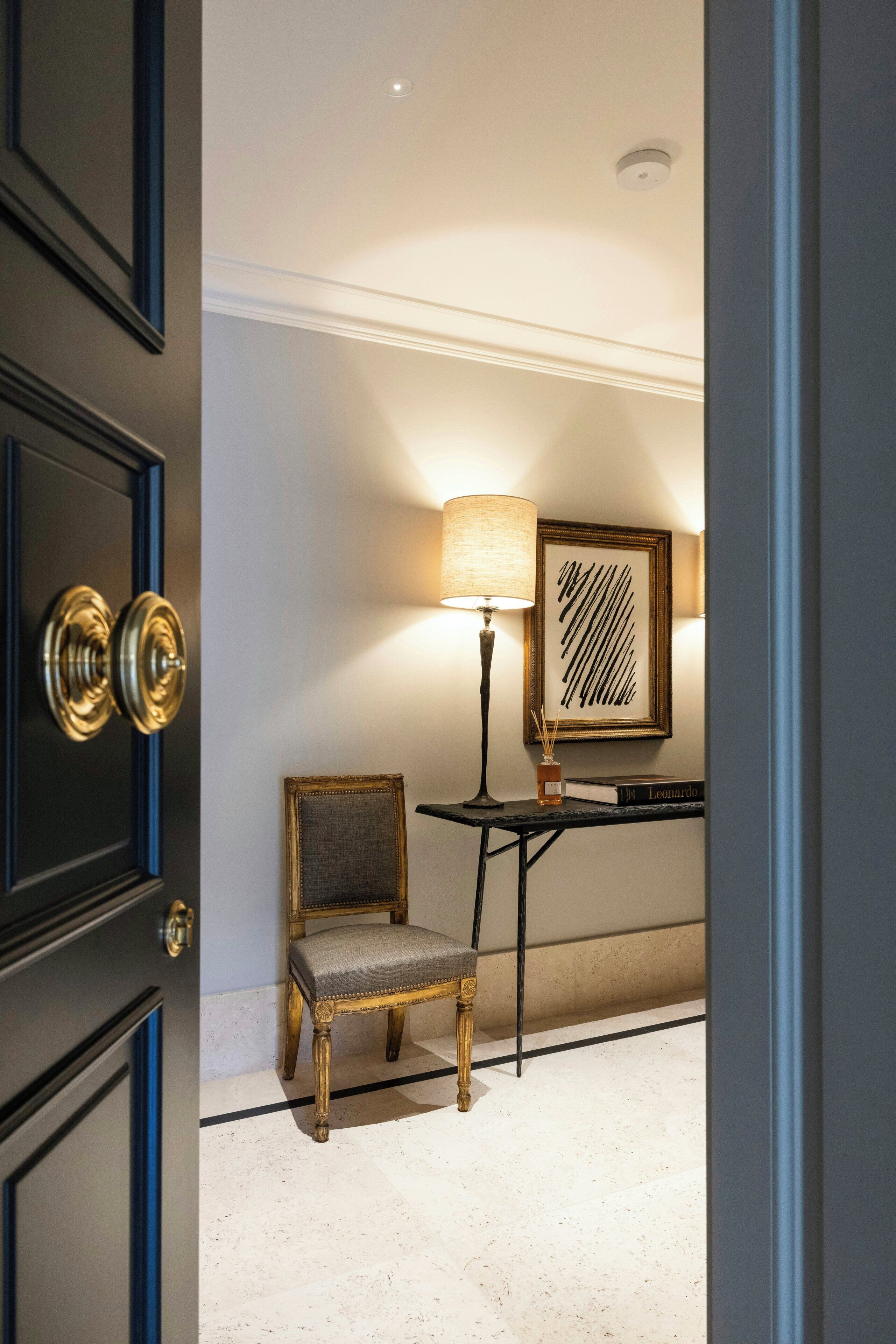 A room with a dark wooden door, a small chair, a table with a lamp, and a framed picture on the wall.