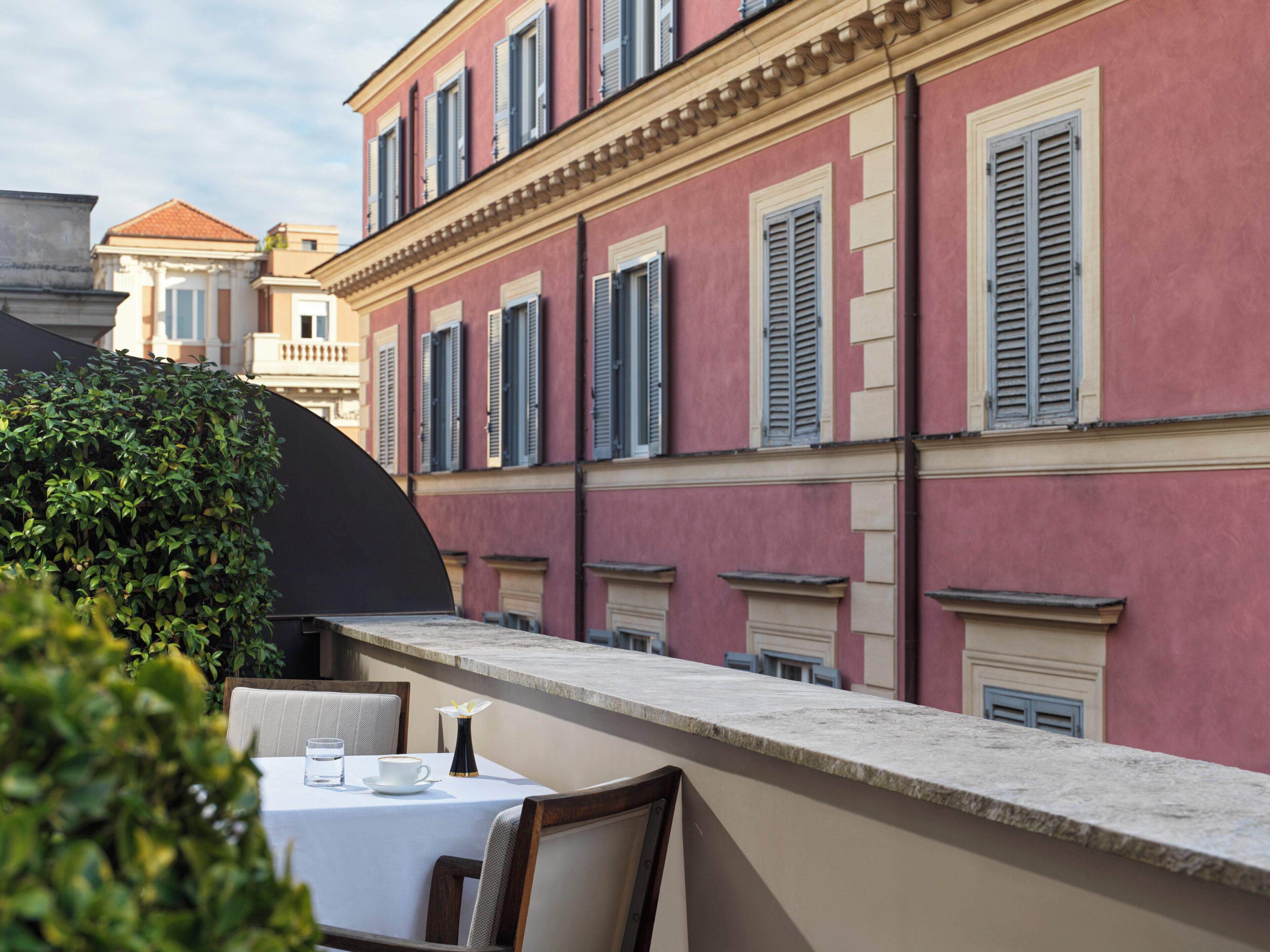 A terrace with a table set for two, overlooking a building with shutters and a balcony.