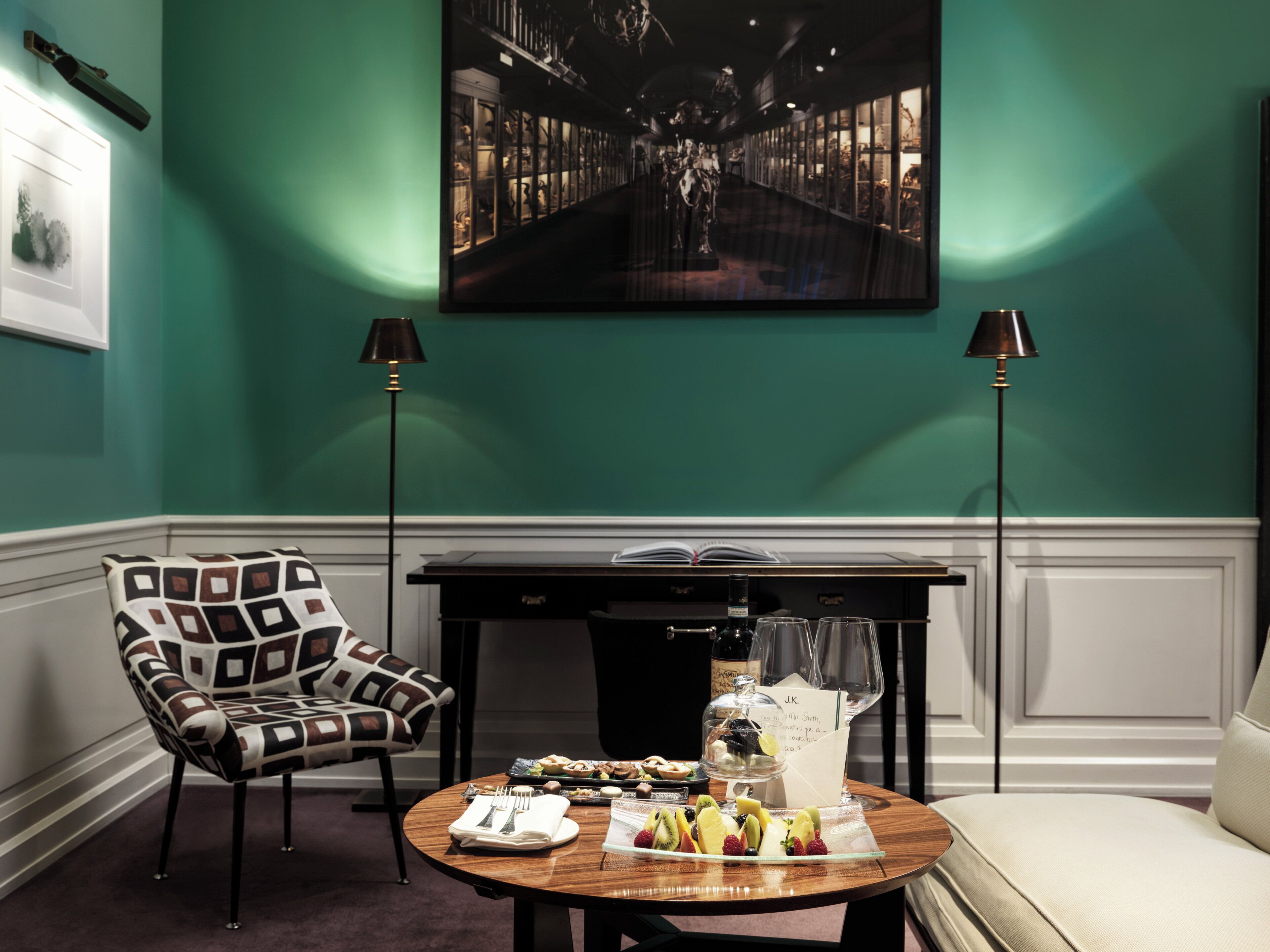 A room with teal walls, a patterned armchair, a dining table with a fruit platter, a bottle, and a menu.