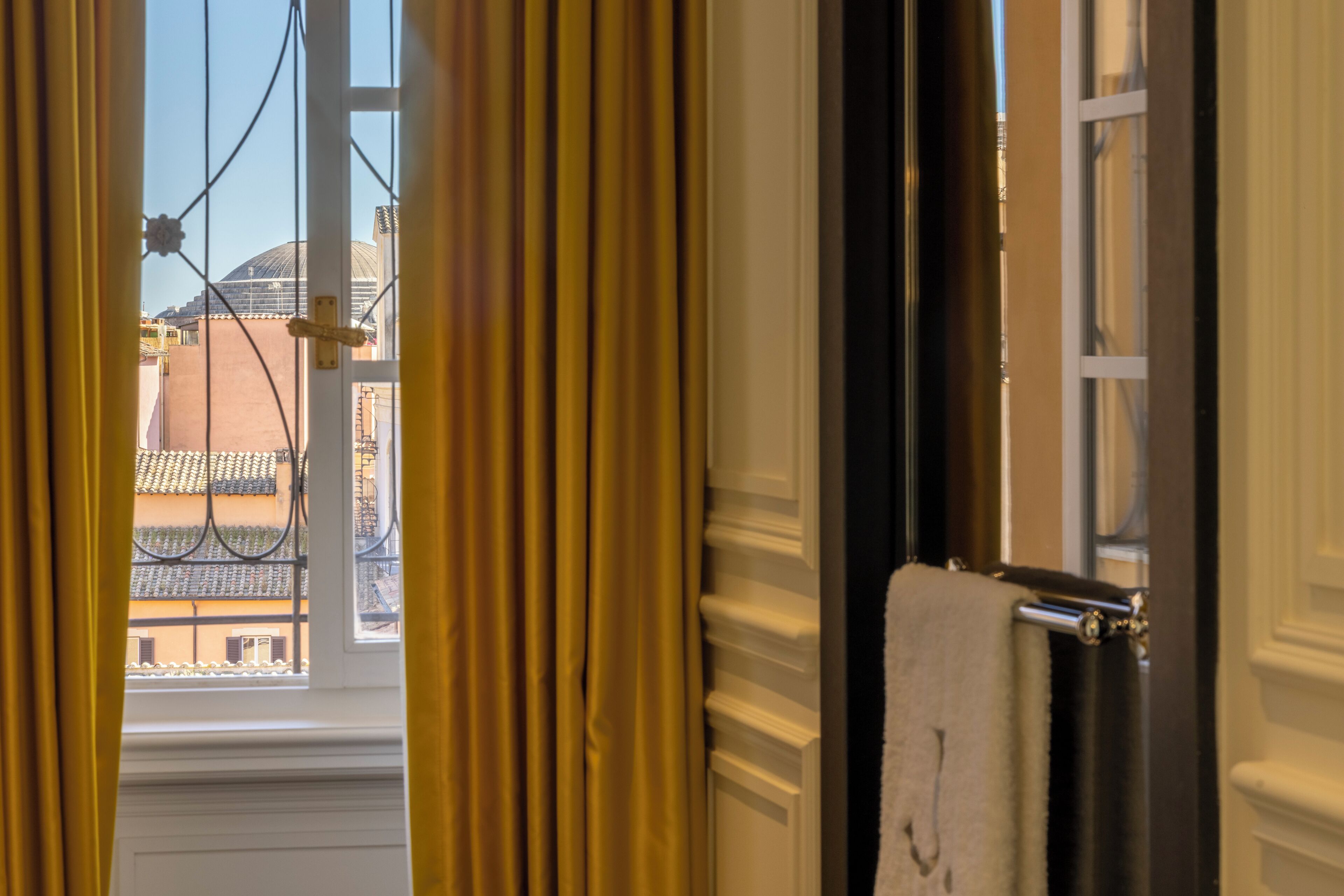 A window with curtains, a towel hanging on a radiator, and a glimpse of a building with a tiled roof.